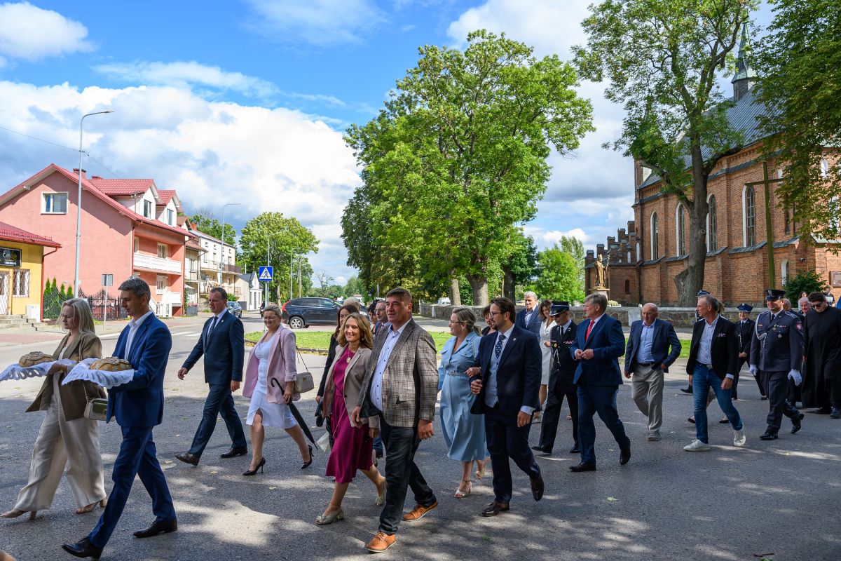 Grupa ludzi w eleganckich strojach idzie ulicą. Tło to zielone drzewa i ceglana zabudowa. Niebo jest błękitne z białymi chmurami.