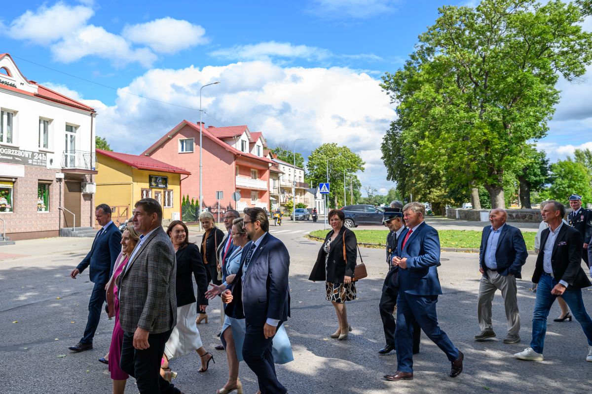 Grupa osób w formalnym ubraniu spaceruje po mieście. Tło to jasne budynki i zielone drzewa pod niebieskim niebem. Atmosfera spotkania w pogodny dzień.