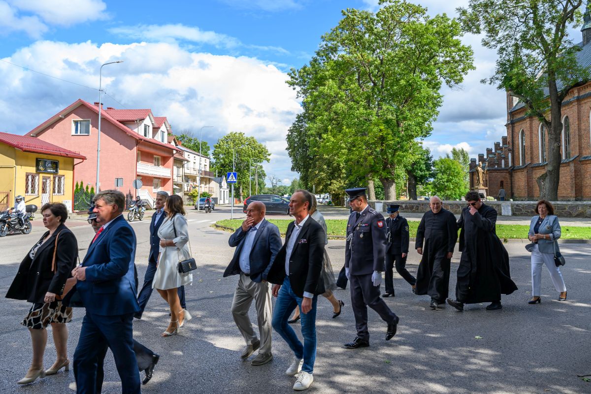 Grupa ludzi spaceruje ulicą w słoneczny dzień. W tle zielone drzewa i ceglana budowla. Po lewej stronie kolorowe domy.