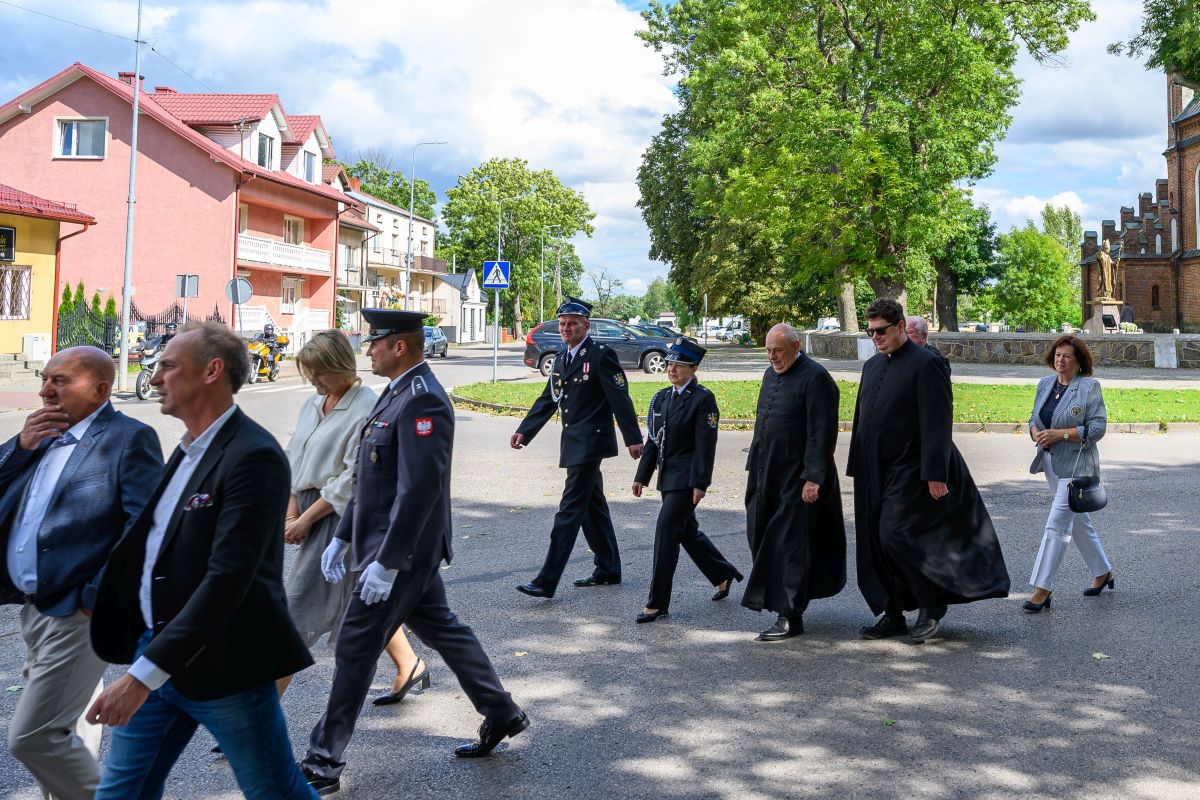 Grupa ludzi w eleganckich ubraniach idzie ulicą. W tle drzewa, czerwony budynek i kościół z cegły. Niebo zachmurzone.