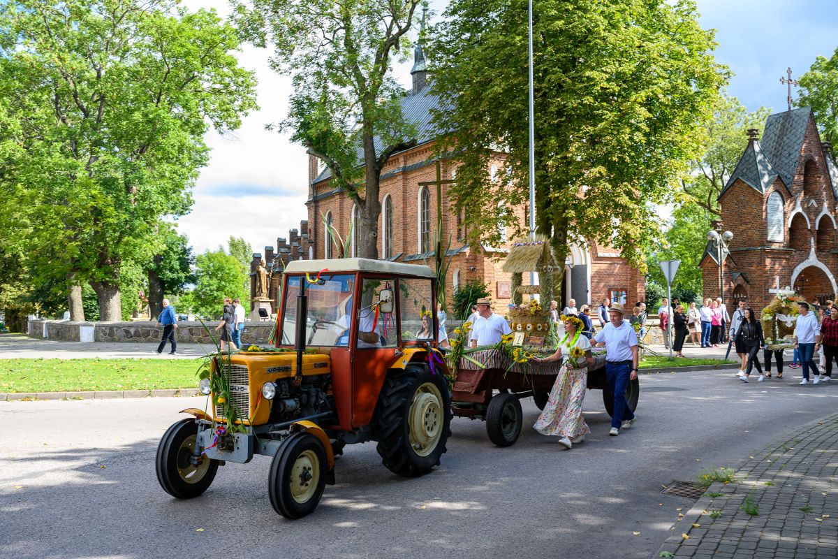 Traktor z ozdobną przyczepą na ulicy, z tyłu ceglana budowla. Ludzie w strojach regionalnych, wokół zielone drzewa.