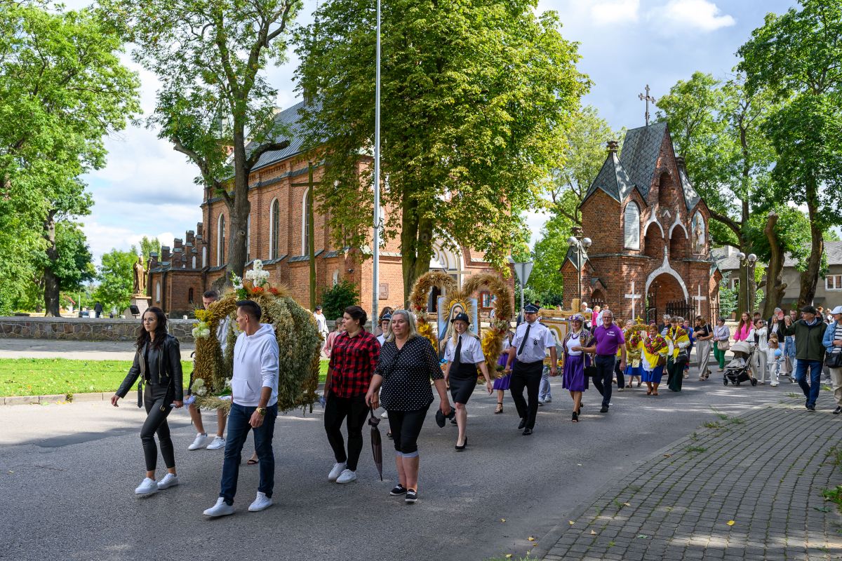 Procesja ludzi idących przed ceglaną świątynią na tle zieleni. Uczestnicy niosą wieńce dożynkowe. Pogodne niebo w tle.