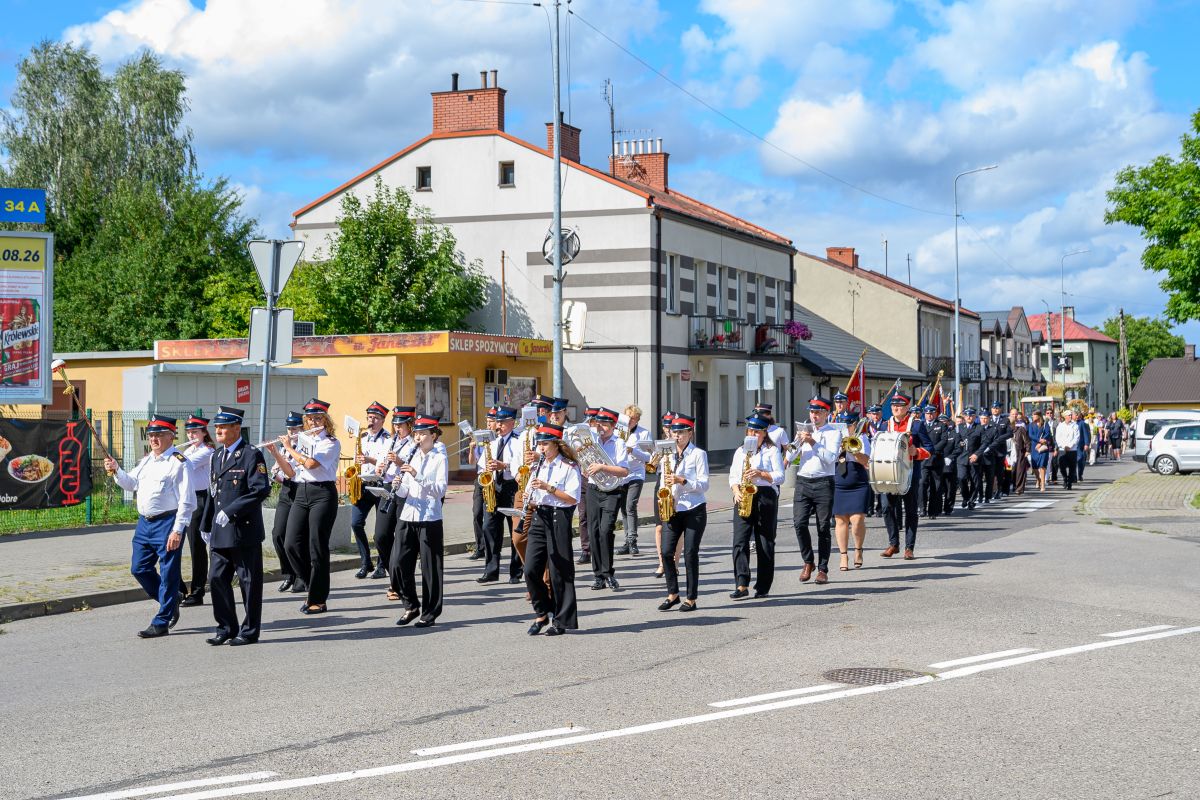 Orkiestra maszeruje ulicą w słoneczny dzień, w tle niskie budynki i drzewa. Uczestnicy w białych koszulach i czapkach. Niebo z chmurami.