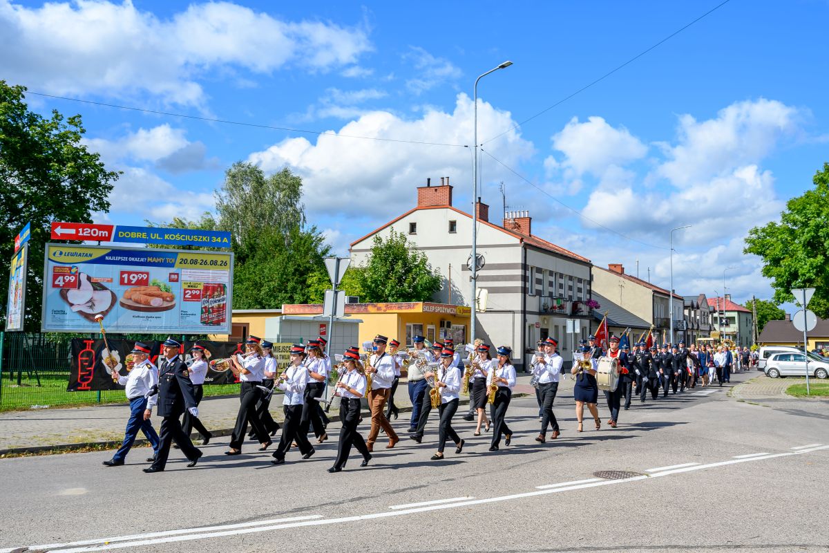 Parada orkiestry dętej na ulicy, w tle budynki i niebo z białymi chmurami. Uczestnicy w uniformach maszerują obok billboardu. Ciepły, słoneczny dzień.