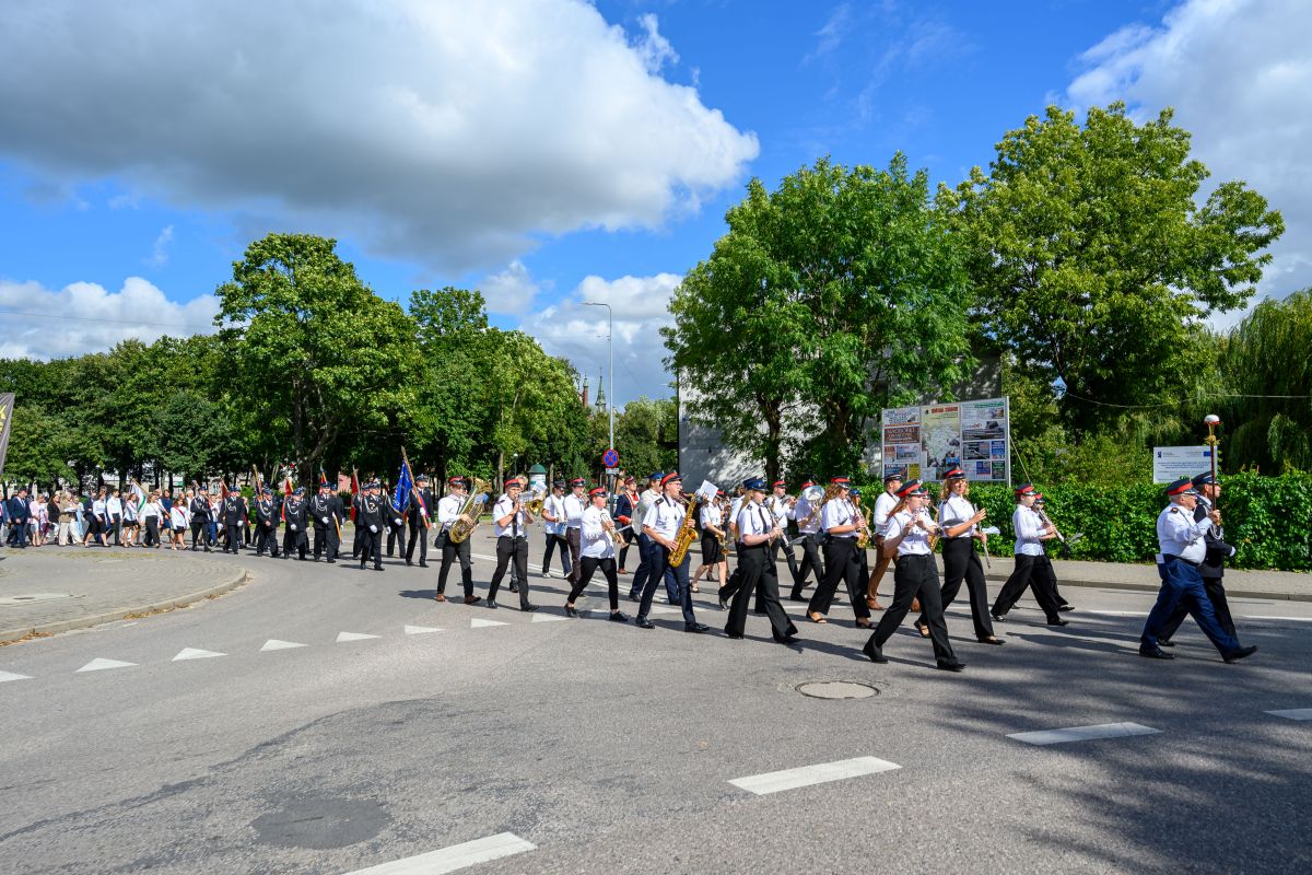 Orkiestra maszeruje ulicą w słoneczny dzień. Muzycy w białych koszulach i czapkach grają na instrumentach dętych. W tle zielone drzewa i niebo z chmurami.