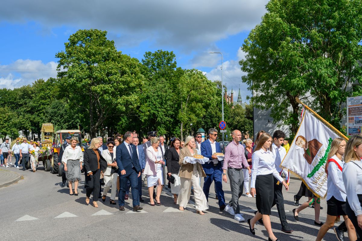 Grupa ludzi na ulicy w słoneczny dzień, trzymają flagi i chleb. Tło stanowią drzewa i niebieskie niebo z chmurami. Uczestnicy są elegancko ubrani.