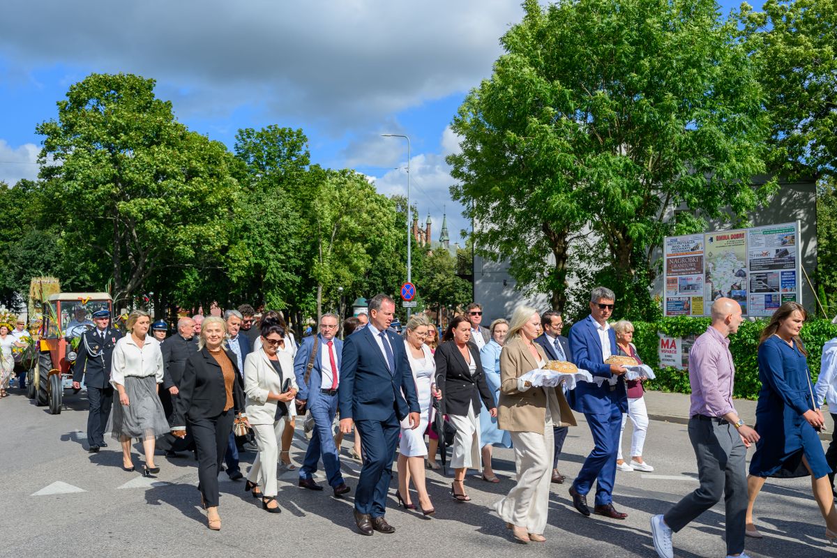 Grupa ludzi ubrana elegancko idzie ulicą, trzymając chleb na białych serwetkach. Tło to zielone drzewa i niebo z chmurami. Po lewej stronie widać traktor z ozdobami.