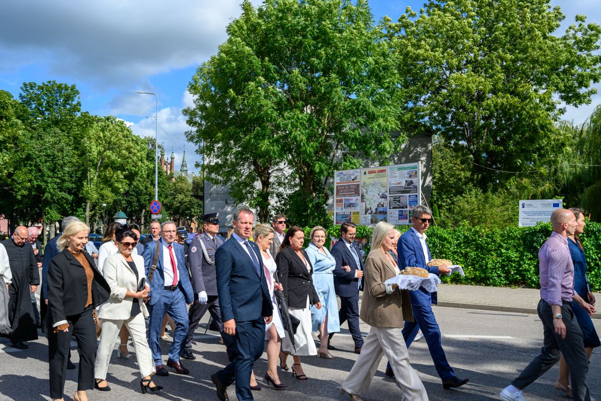 Grupa ludzi w eleganckiej odzieży idzie ulicą na tle zielonych drzew. Jasne niebo z chmurami, po prawej strony tablice ogłoszeniowe. Osoba niesie tradycyjny chleb na białym obrusie.