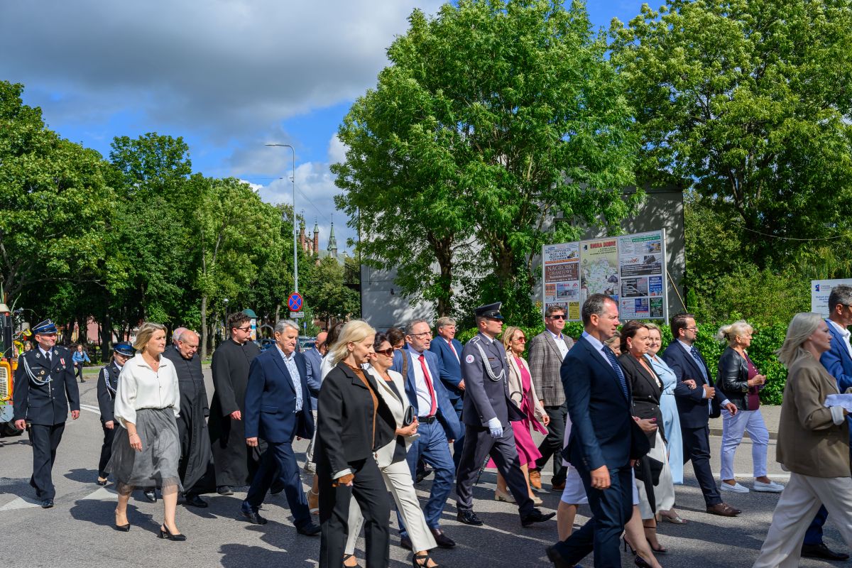 Grupa ludzi w eleganckich strojach i mundurach idzie ulicą. Tło to zieleń drzew, niebo częściowo pochmurne. Po prawej widoczne są plakaty.