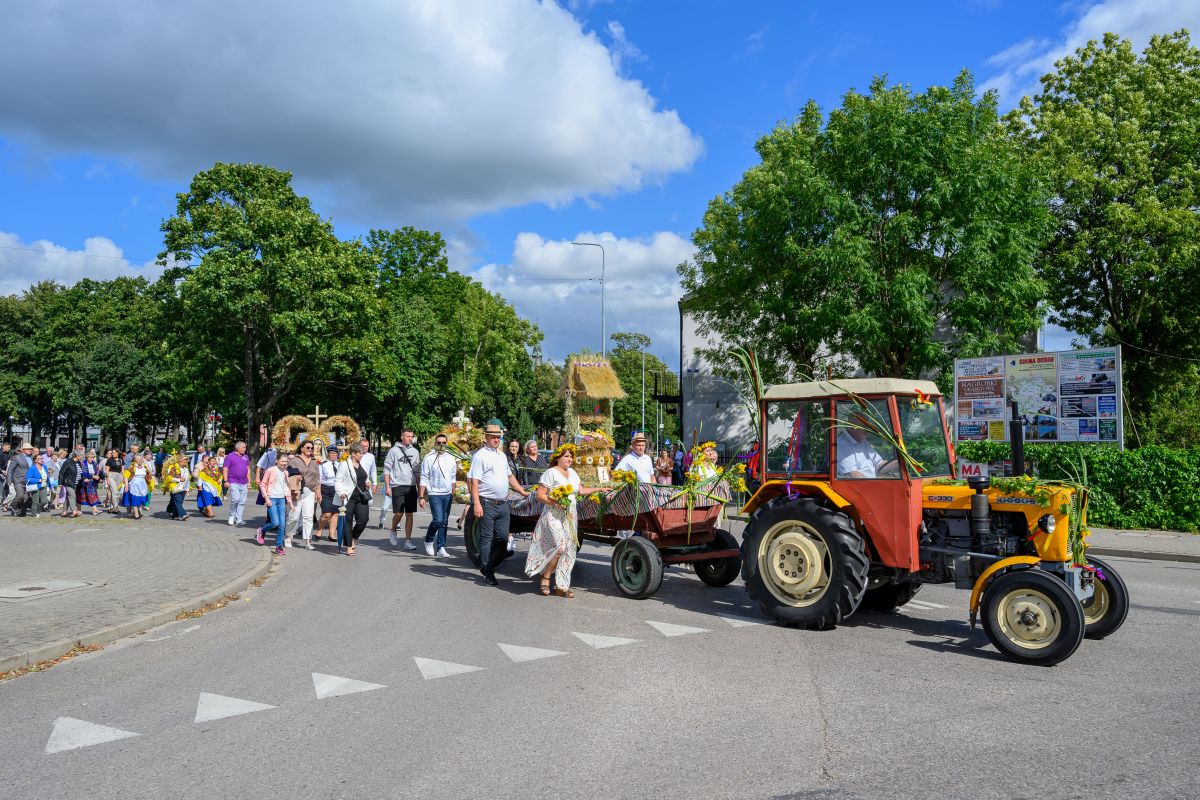 Grupa ludzi w tradycyjnych strojach idzie za czerwonym traktorem ciągnącym przyczepę z kwiatami. Tło z zielonymi drzewami i niebieskim niebem. Spokojna scena festiwalowa na ulicy.