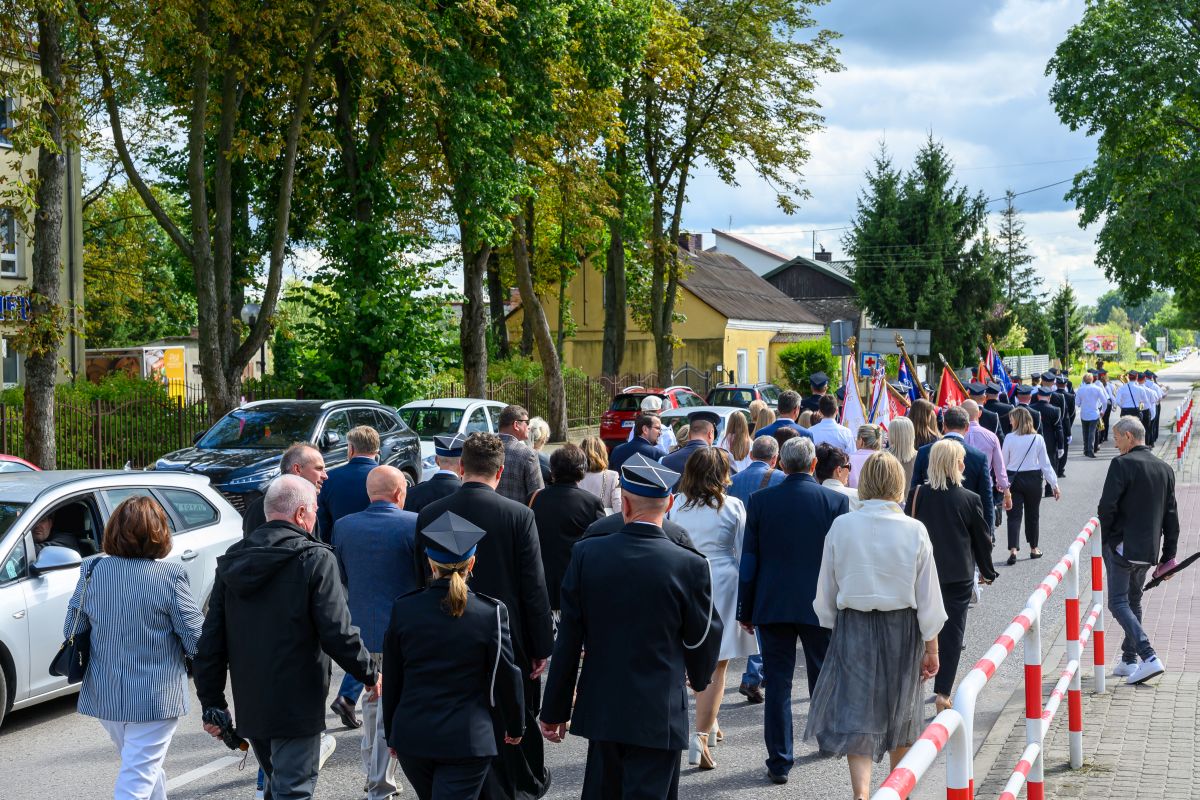 Grupa ludzi w eleganckich ubraniach idzie ulicą w uroczystym pochodzie. Na tle zielonych drzew widoczne są kolorowe flagi. Pogodne niebo z białymi chmurami.