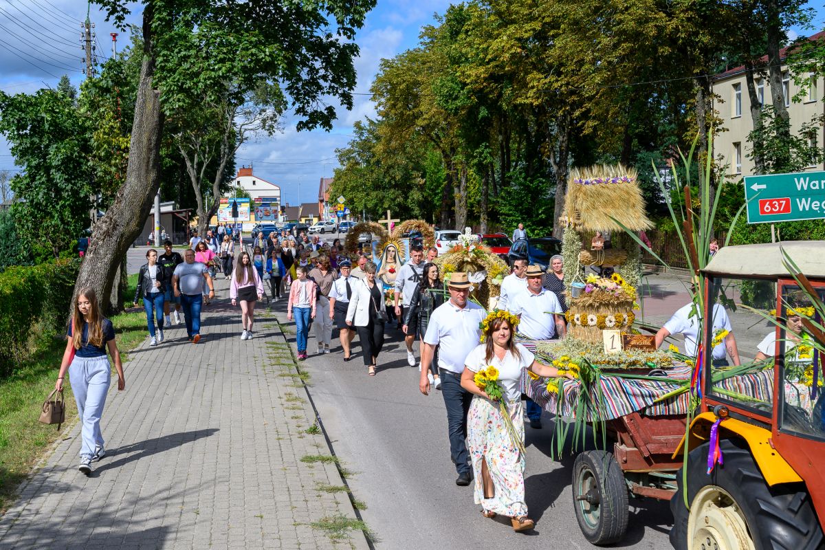Parada dożynkowa na drodze, ludzie w odświętnych strojach, kolorowe dekoracje. Drzewa w tle, jasno i słonecznie. Traktor z wieńcem z przodu.
