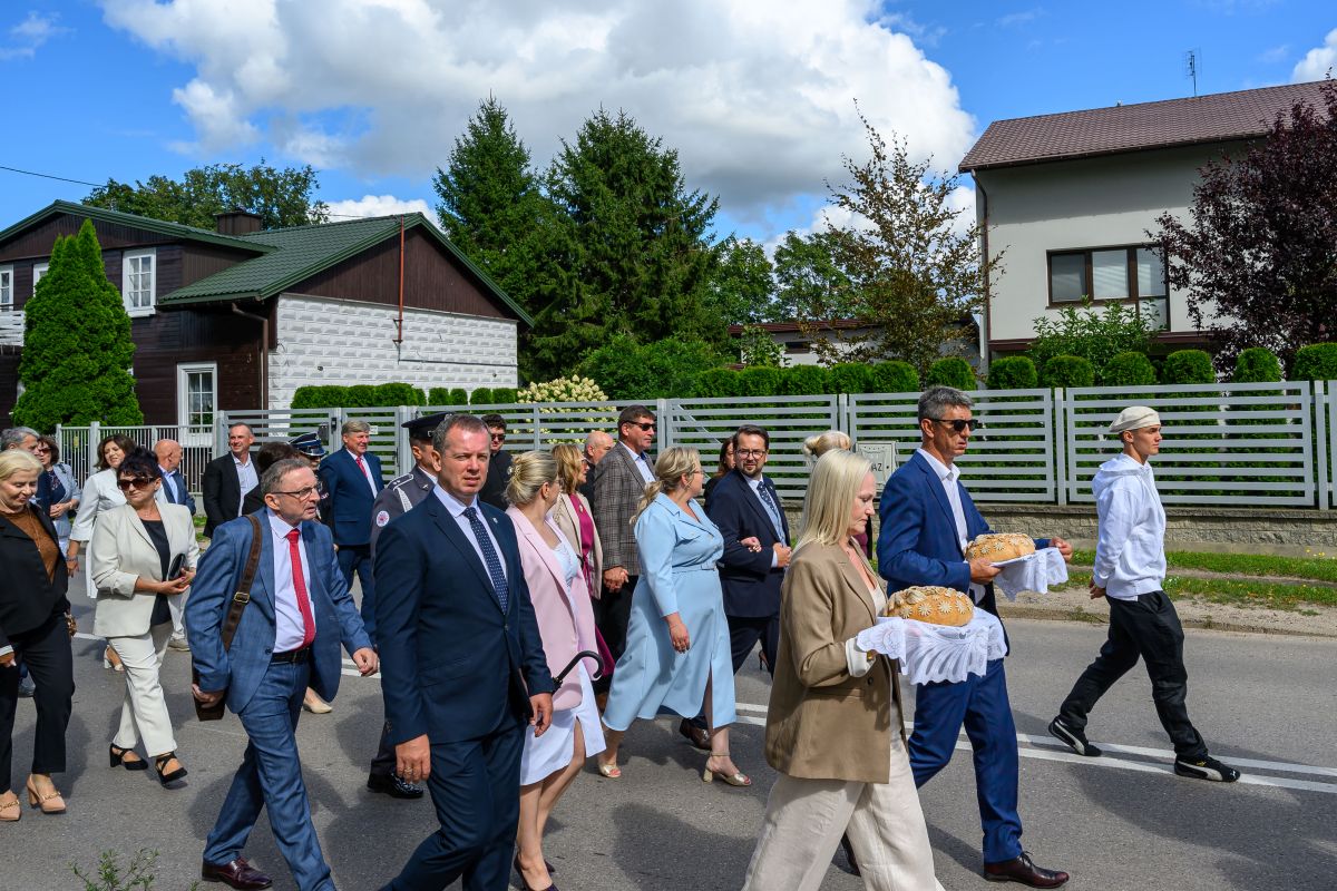 Grupa elegancko ubranych ludzi idzie ulicą, niosąc chleb na białym obrusie. Tło to domy i zielone drzewa. Pogoda słoneczna, niebo z chmurami.