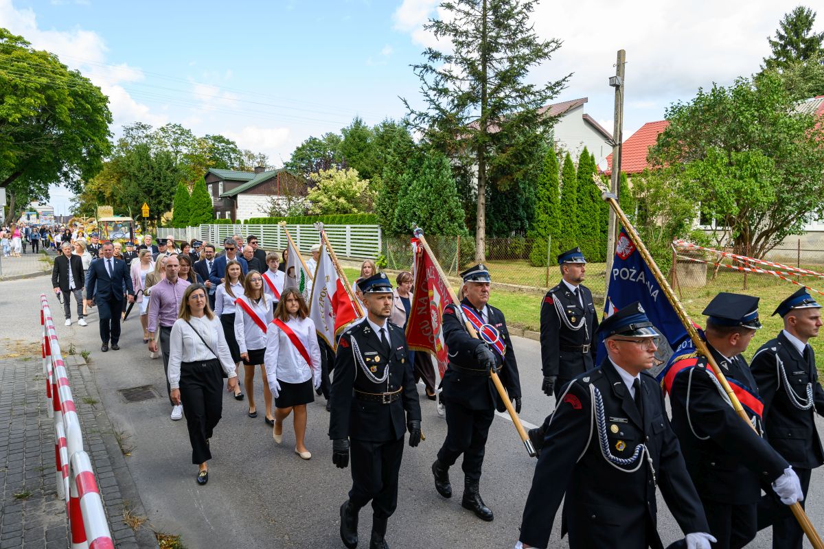 Na zdjęciu widoczny jest pochód strażaków w czarnych mundurach z chorągwiami. W tle znajdują się zielone drzewa i domy z czerwonymi dachami. Na niebie widać kilka białych chmur.