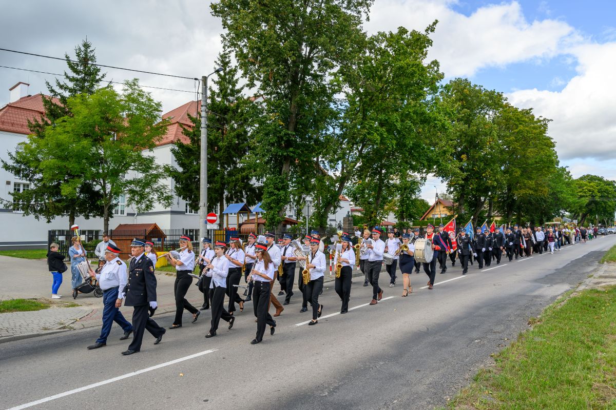 Grupa muzyków maszeruje wzdłuż ulicy z drzewami w tle. Ubrani w białe koszule i czarne spodnie grają na instrumentach dętych. W tle widać zielone drzewa i budynek z czerwonym dachem.