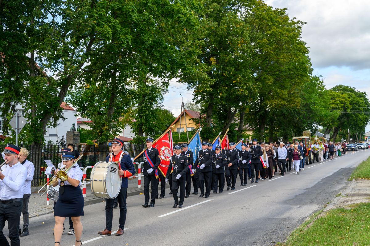 Parada na ulicy z ludźmi w mundurach, orkiestra z instrumentami i kolorowe sztandary. Drzewa w tle, zielone liście. Pogoda pochmurna.