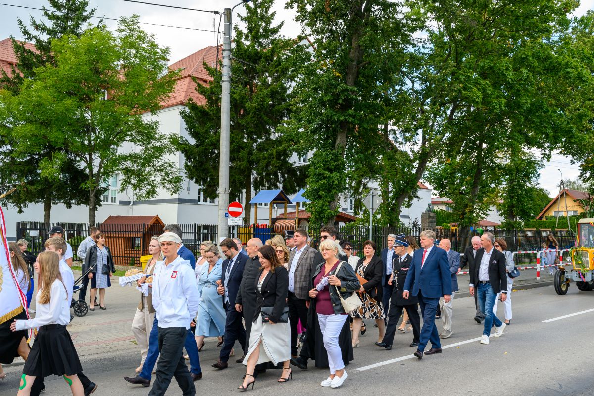 Grupa ludzi spaceruje ulicą przy zielonych drzewach i budynku z czerwonym dachem. Mają eleganckie stroje. Tło ożywia plac zabaw.