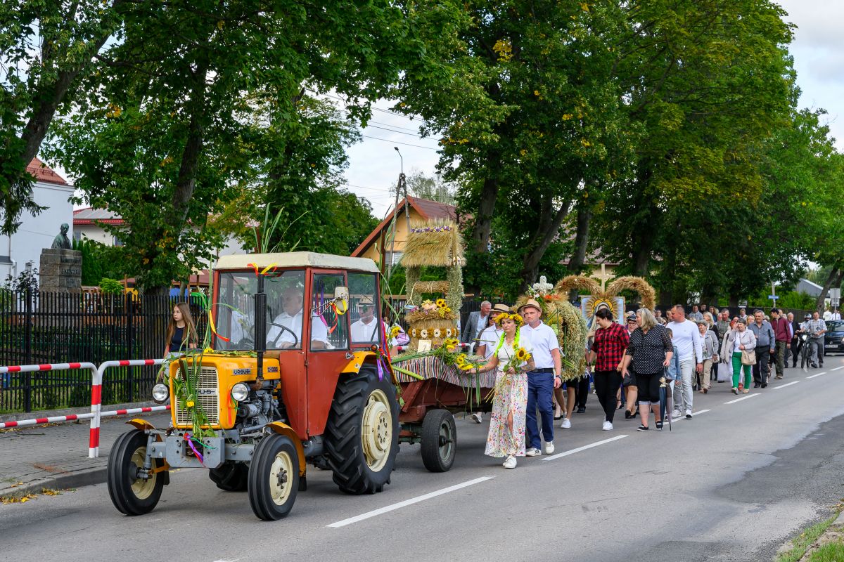 Parada z traktorem w wiejskim stylu, kolorowy wóz z kwiatami, obok tłum ludzi, zielone drzewa w tle.