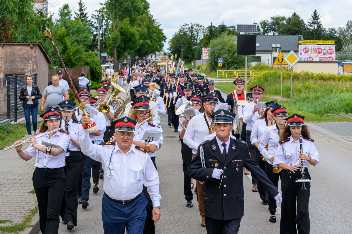 Orkiestra dęta maszeruje ulicą w białych koszulach i czapkach, grając instrumenty. W tle zielone drzewa i znaki drogowe. Okolica jest miejska z niską zabudową.
