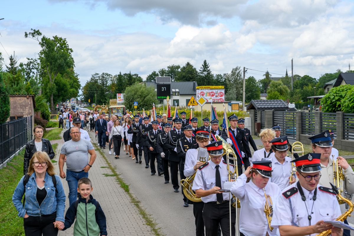 Pochód orkiestry dętej i strażaków w mundurach idących ulicą. Domy i zieleń w tle, jasne niebo z chmurami. Uczestnicy niosą instrumenty i flagi.