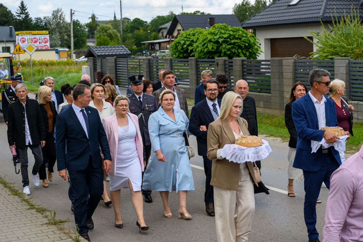 Grupa ludzi w eleganckich strojach idzie ulicą. Kobieta i mężczyzna niosą chleby na białych serwetkach. W tle zielone ogrody i domy.