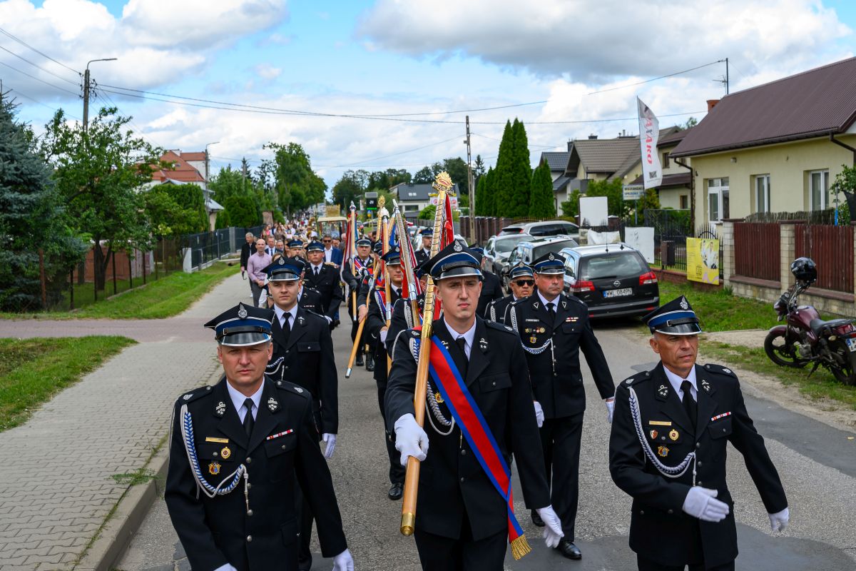 Parada strażaków w czarnych mundurach z odznaczeniami i sztandarami na wiejskiej ulicy. Tło stanowią domy i drzewa. Niebieskie niebo z chmurami.
