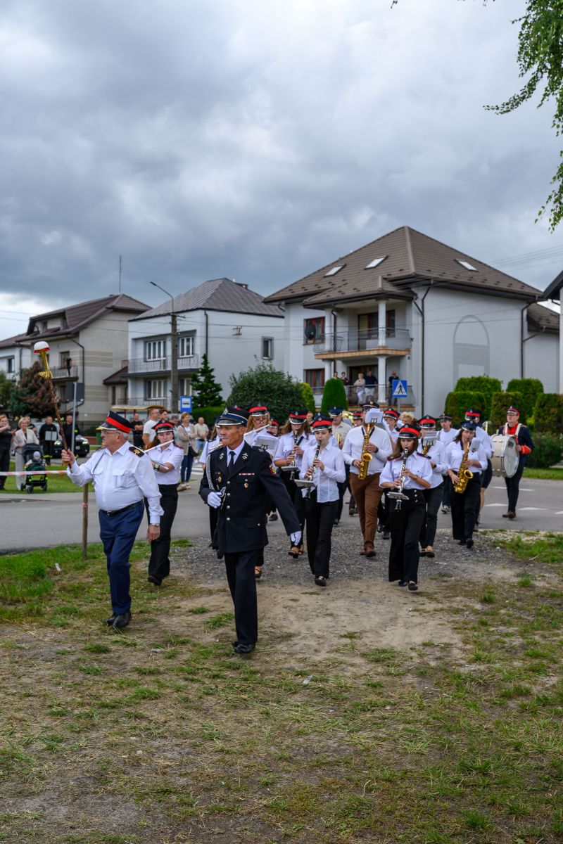 Orkiestra strażacka w białych i czarnych strojach maszeruje ulicą. W tle są domy i zachmurzone niebo. Towarzyszy im grupa mieszkańców.