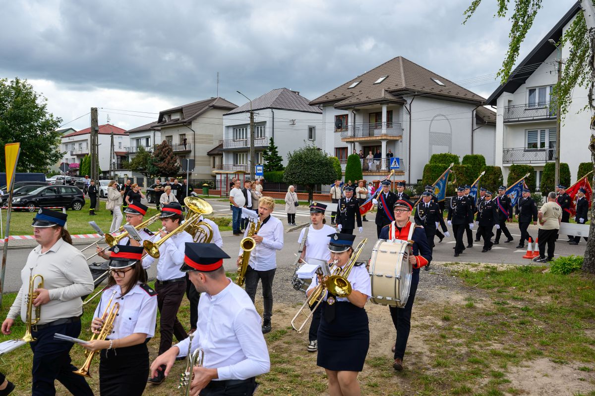 Orkiestra maszeruje ulicą przed białymi budynkami. Ubrania muzyków w bieli, czerni i czerwieni. Tło to chmury i widzowie.