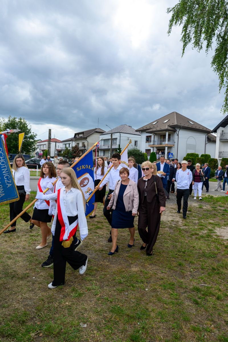 Grupa młodzieży w białych koszulach niesie sztandary na tle domów i chmur. Tło jest zielone i pochmurne. Eleganckie ubrania wskazują na uroczystość.