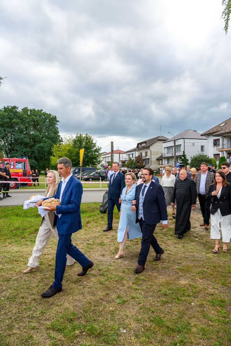 Grupa ludzi idzie po trawiastej ścieżce; na czele mężczyzna niesie chleb. W tle stoją domy i czerwony wóz strażacki. Pochmurne niebo.