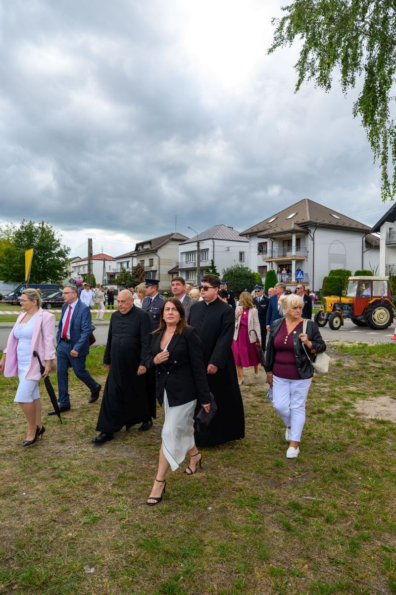 Grupa ludzi w formalnych strojach idzie wzdłuż trawiastej drogi. W tle widać budynki mieszkalne i traktor. Niebo jest zachmurzone.