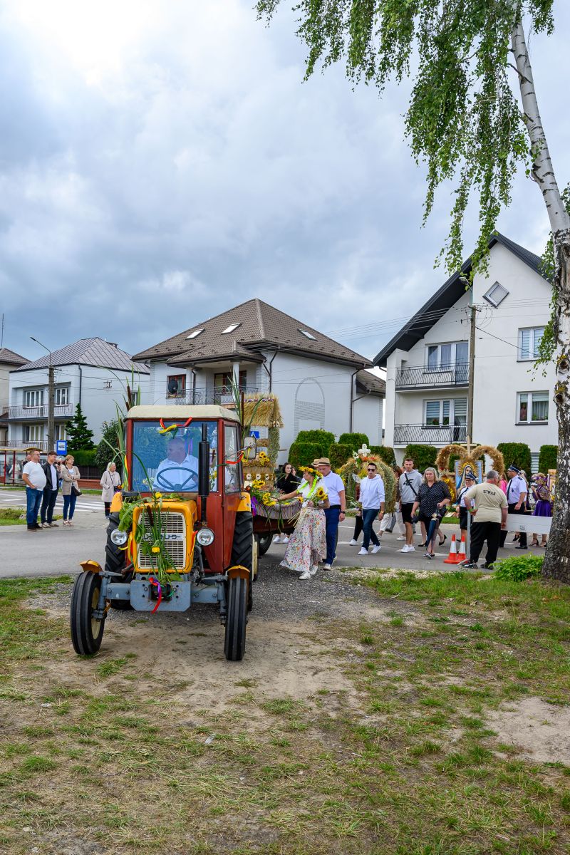 Na zdjęciu widać kolorowy traktor z przodu, ozdobiony wstążkami i kwiatami. W tle są ludzie oraz białe domy z szarymi dachami. Niebo jest zachmurzone, a po prawej stoi brzoza.