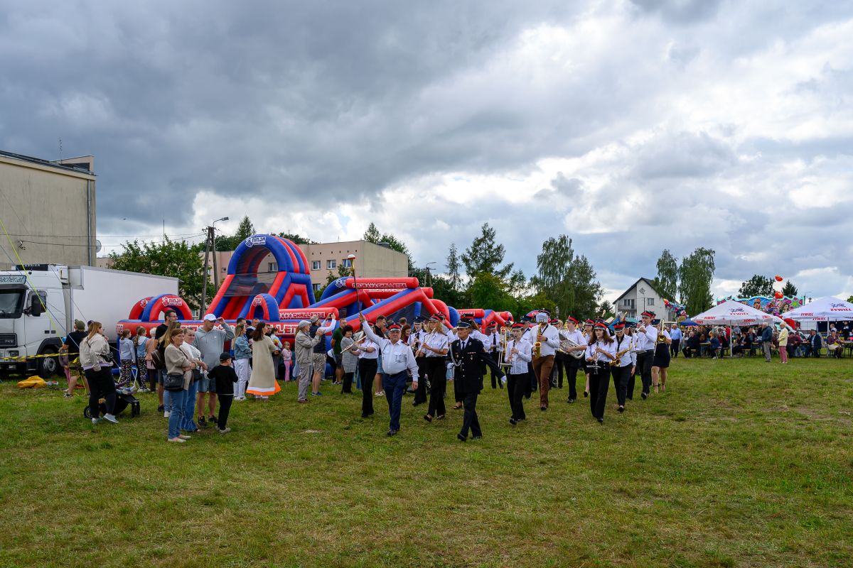 Marsz orkiestry na tle kolorowego dmuchanego placu zabaw oraz zielona polana. Obok widoczni są ludzie. Niebo pochmurne.