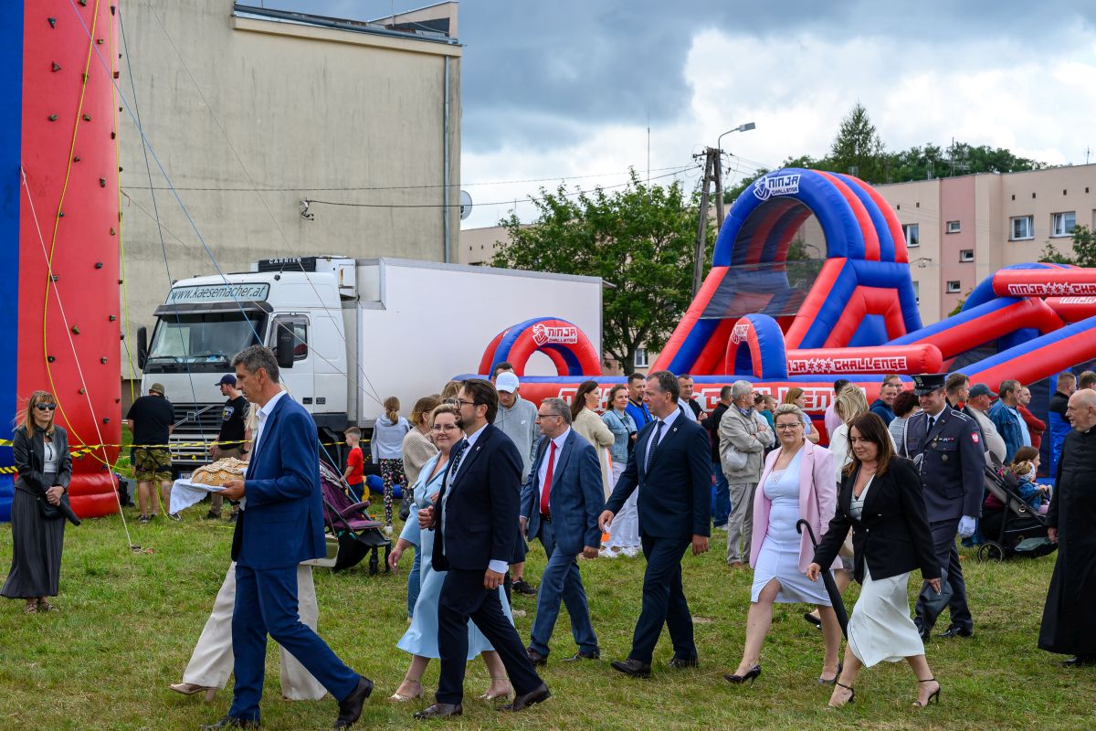 Grupa ludzi w eleganckich strojach idzie po trawie na tle kolorowego dmuchanego toru przeszkód. Nad nimi chmury. W tle widoczny biały samochód dostawczy.