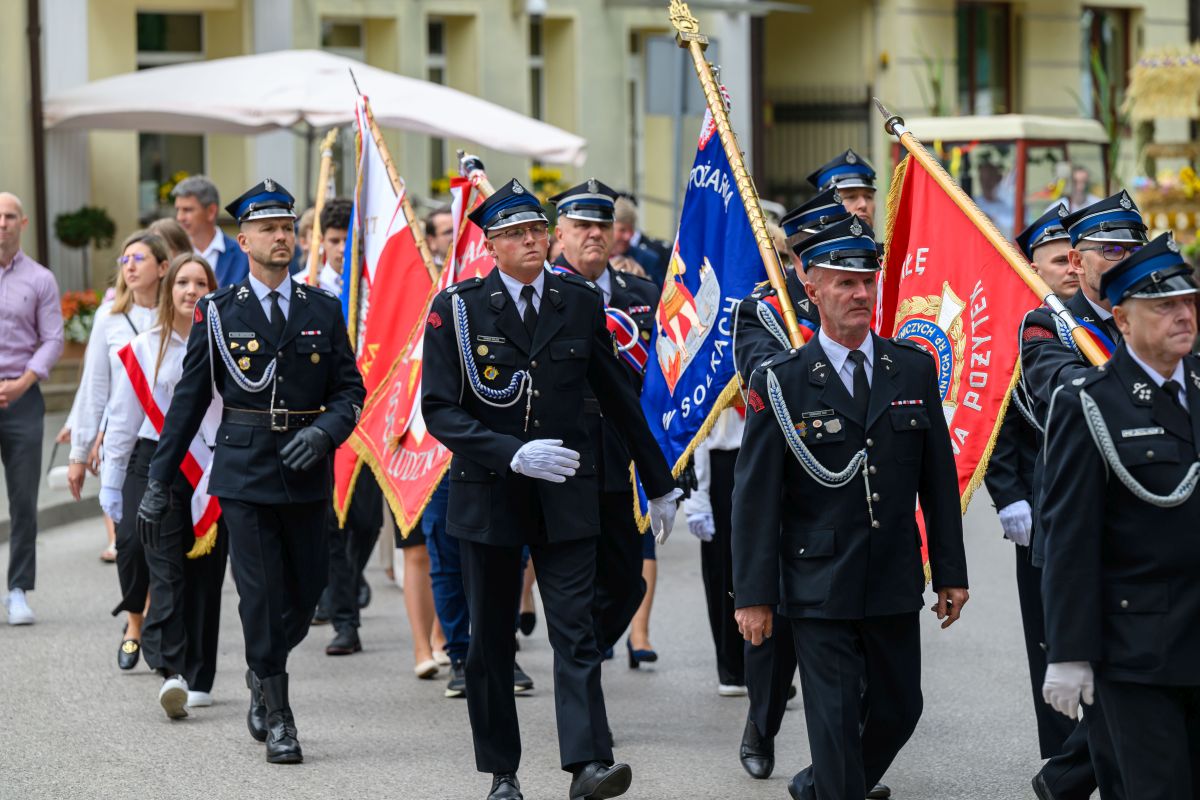 Grupa strażaków w czarnych mundurach maszeruje z kolorowymi sztandarami. W tle budynek i ludzie. Dominuje czerń i czerwień.