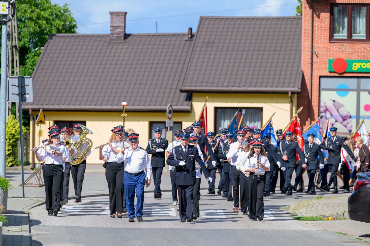 Parada orkiestry dętej w mundurach maszeruje ulicą. Jasne, letnie słońce, kolorowe flagi w tle. Domy w tle mają dachy w odcieniach brązu.