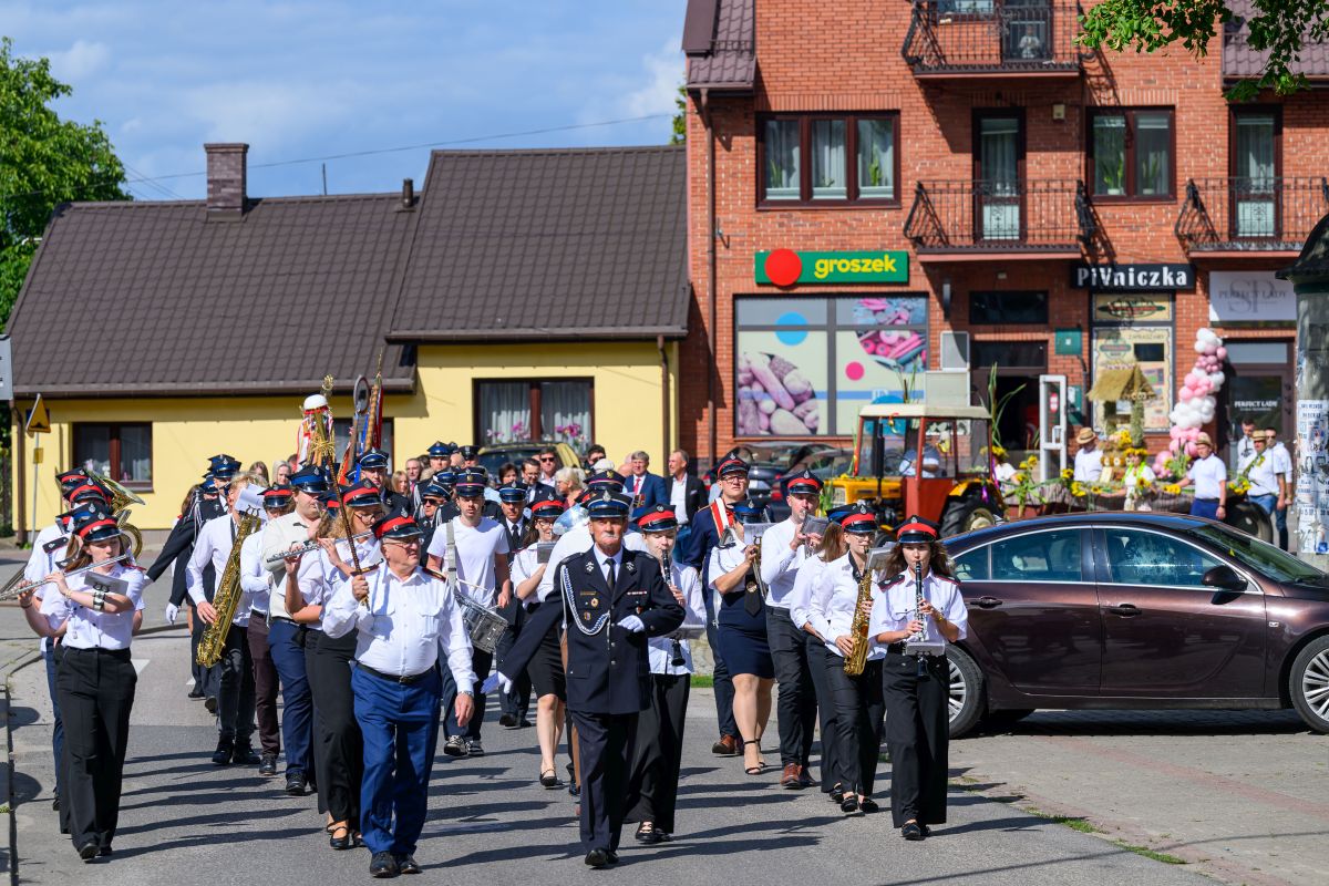 Orkiestra dęta maszeruje ulicą w słoneczny dzień. Członkowie noszą mundury w odcieniach bieli, granatu i czerwieni. W tle żółty budynek i czerwony sklep groszek.
