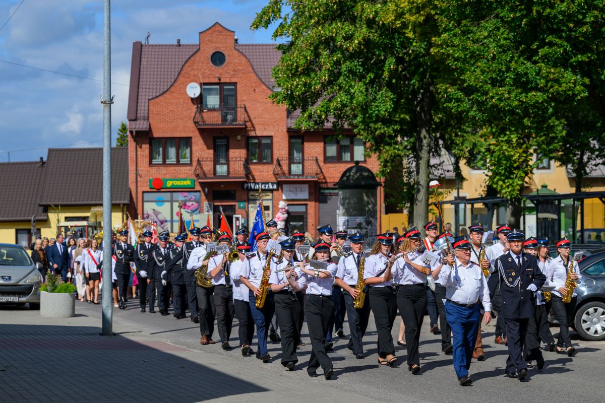 Parada strażaków w mundurach i orkiestrą maszeruje ulicą. Czerwony budynek w tle, zielone drzewa po prawej. Słoneczna pogoda.