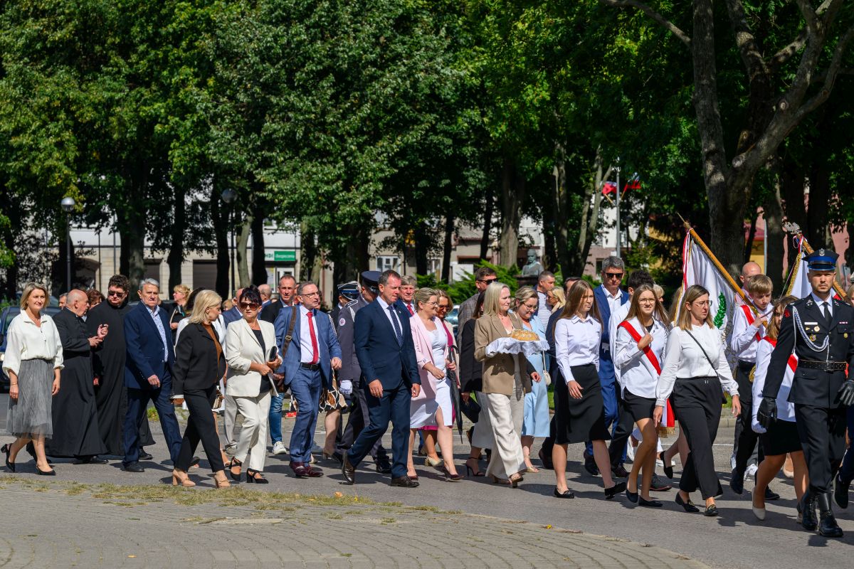 Grupa ludzi na paradzie, ubrani elegancko, niosą flagi i chleb. Tło zielone drzewa, słoneczny dzień. Uroczysta atmosfera.