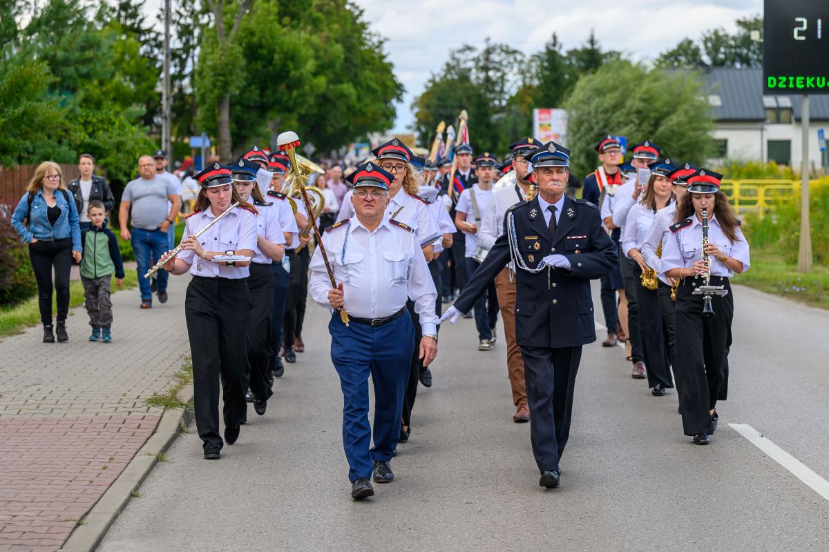 Orkiestra strażacka maszeruje ulicą, ubrane w biało-niebieskie mundury. Tło to zielone drzewa i kilka widzów. Instrumenty to trąbki i klarnety.