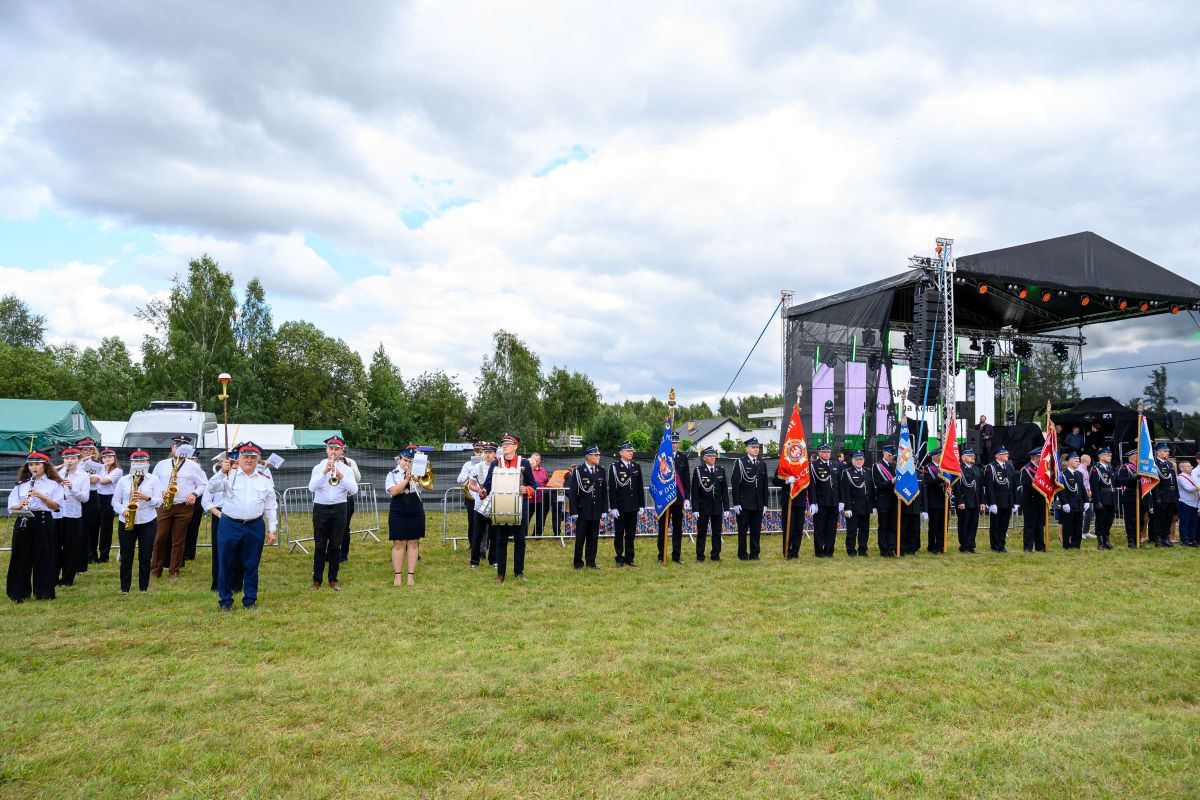 Sześcioosobowy zespół dęty i parada honorowa na zielonej trawie. W tle scena i drzewa. Niebo pochmurne.