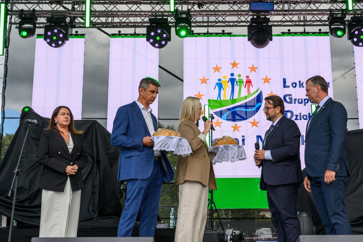 Pięć osób stoi na scenie, wręczając chleb. Tło stanowi ekran z kolorowym logo. Dominują odcienie niebieskiego i zielonego.