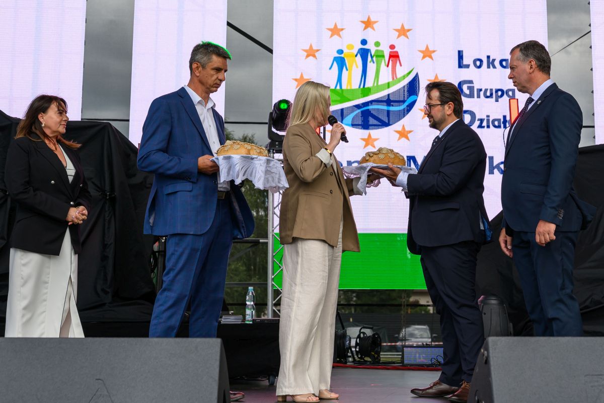 Grupa osób w eleganckich strojach na scenie wymienia się chlebem. Tło to jasny ekran z kolorowym logo. Atmosfera oficjalna.
