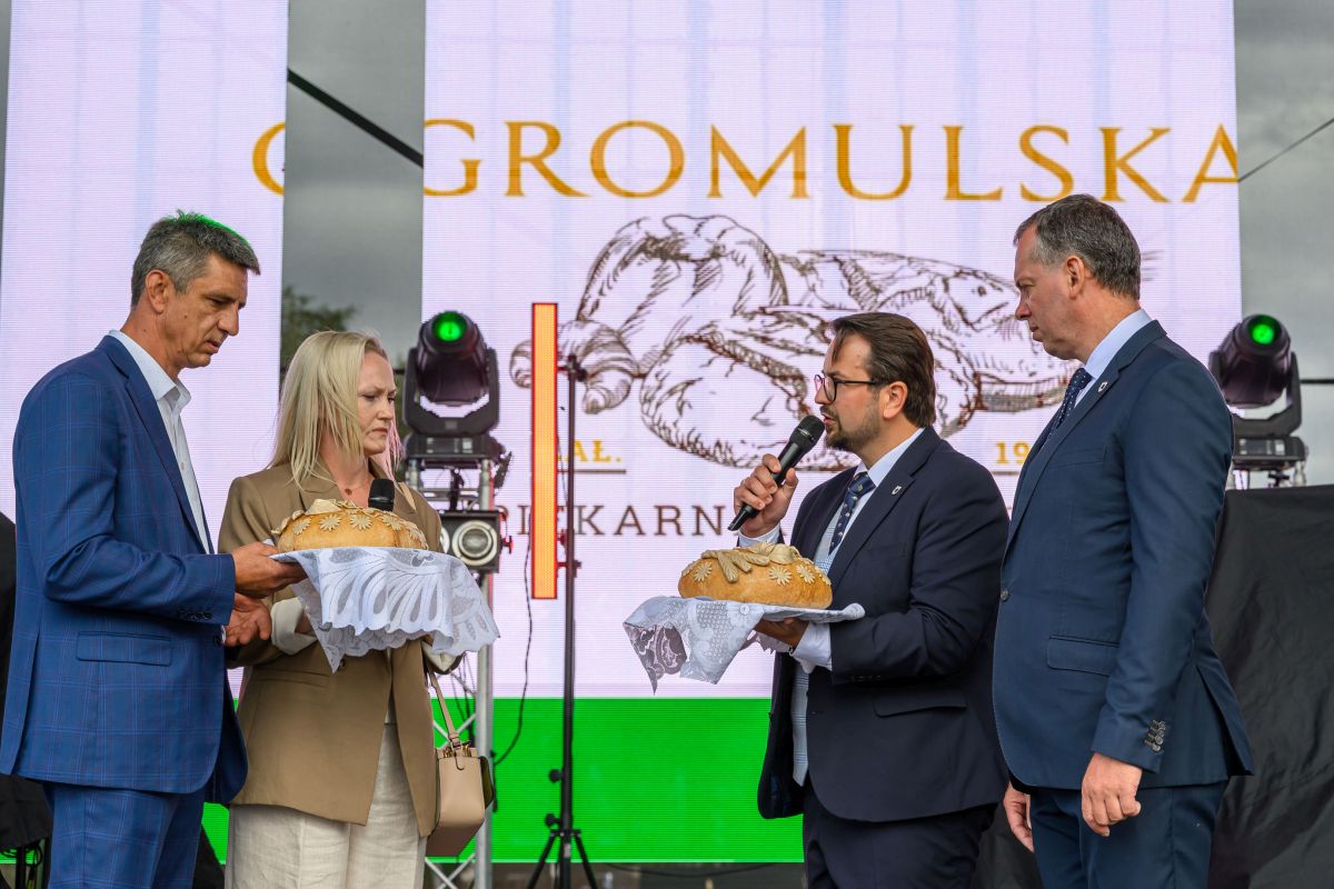 Cztery osoby na scenie prezentują tradycyjne chleby na białych serwetach. W tle widoczny jest baner z napisem „Gromulska”. Sceneria jest kolorowa i uroczysta.