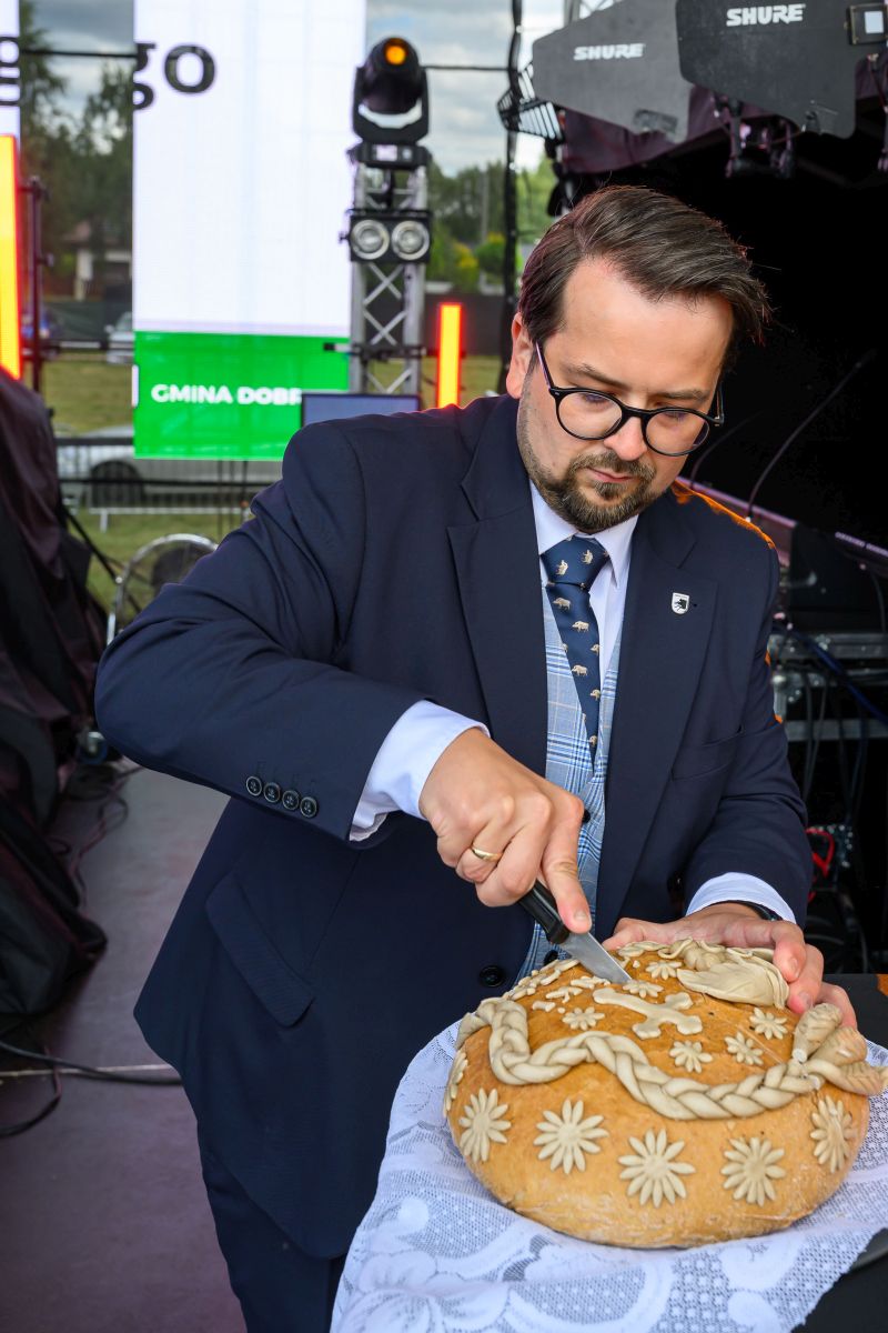 Mężczyzna w garniturze kroi ozdobny chleb. Tło stanowią kolorowe światła i zielona tablica. Ujęcie sceniczne na świeżym powietrzu.