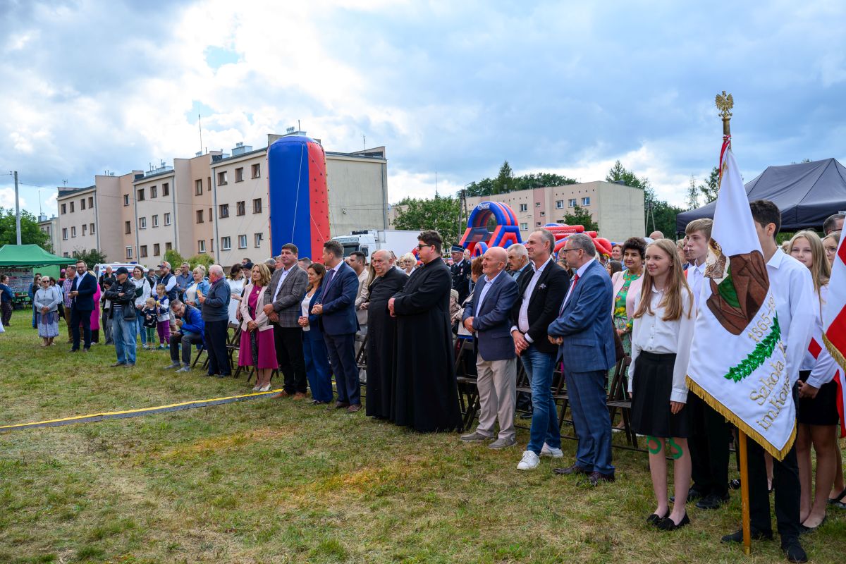 Grupa ludzi uczestniczy w plenerowym wydarzeniu. W tle widoczne są budynki i kolorowe dmuchańce. Niebo jest pochmurne.