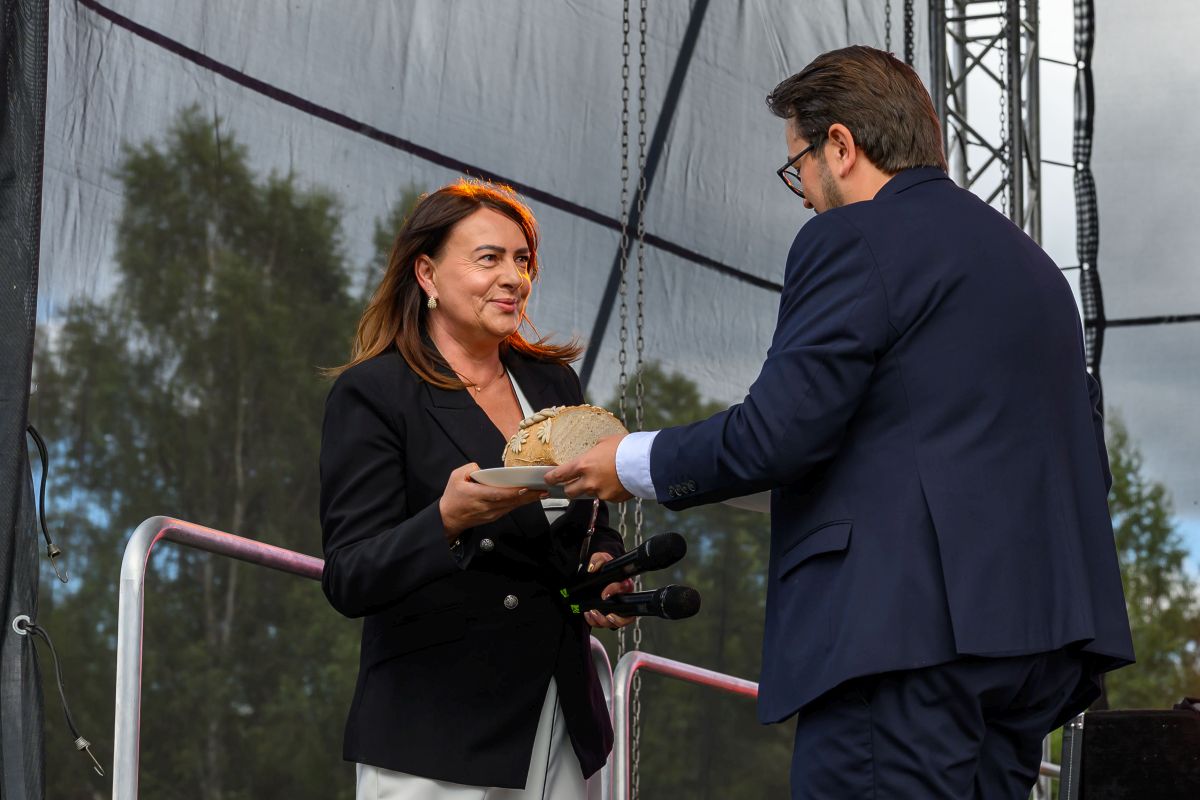 Kobieta w czarnym żakiecie trzyma mikrofony, mężczyzna podaje jej chleb na białym talerzu. Tło sceniczne z drzewami.