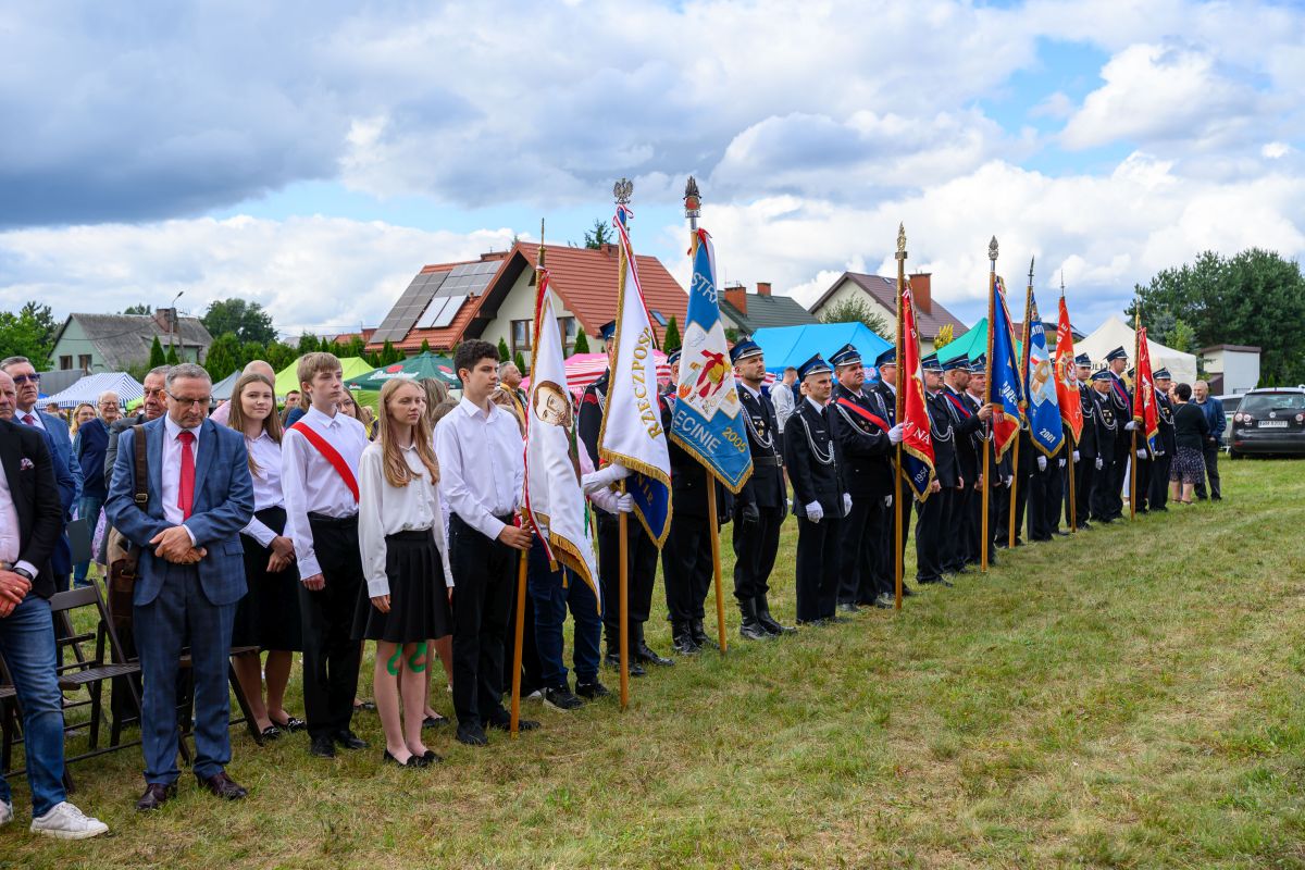 Grupa ludzi z flagami stoi na trawie, ubrani są odświętnie. W tle domy i namioty, niebo pokryte chmurami. Kolory flag są różnorodne.