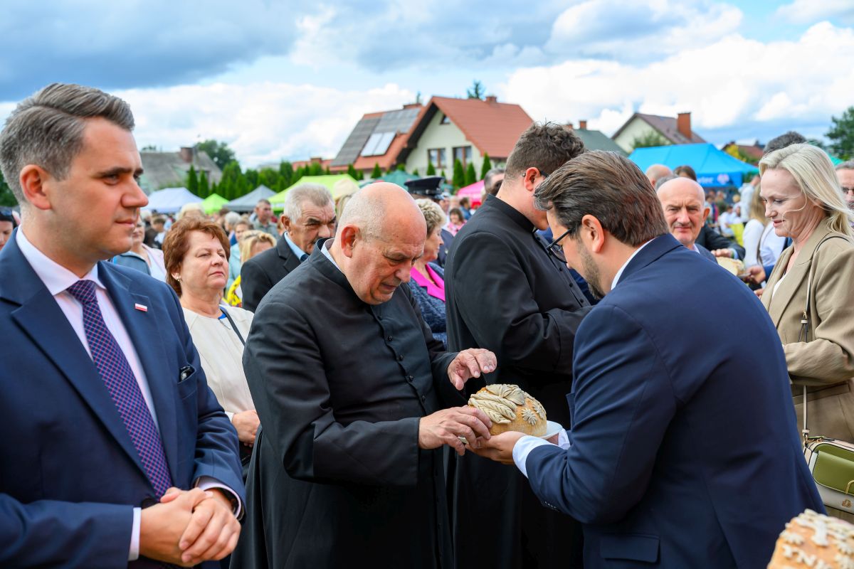 Grupa ludzi na zewnętrznej ceremonii, jasne niebo z chmurami. Kapłan w czarnej sutannie odbiera chleb. Tło z zielonymi namiotami i domami.