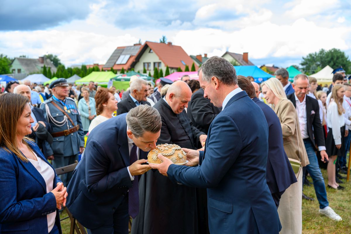 Na obrazie widać uroczystość plenerową z udziałem wielu osób. Mężczyzna całuje tradycyjny chleb, trzymany przez innego mężczyznę. W tle namioty i zachmurzone niebo.